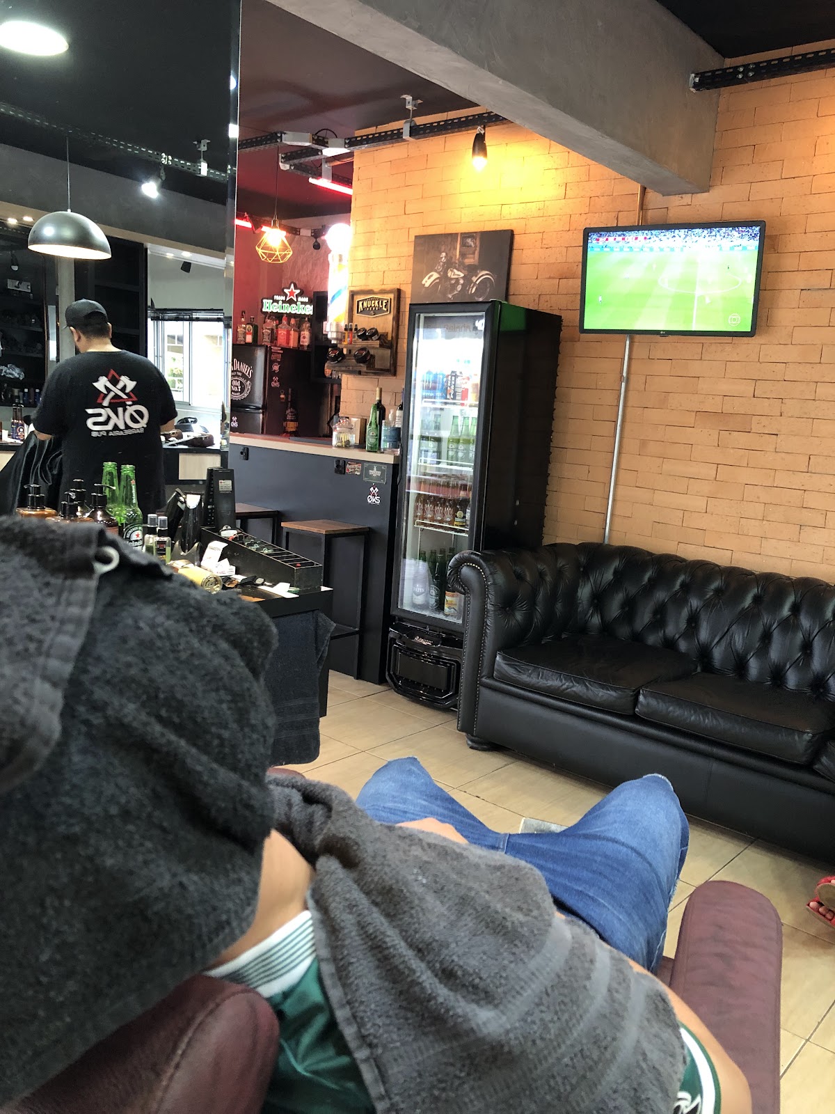 Área de bar da Øks Barbearia - Pub, com freezer de cerveja, sofá e televisão